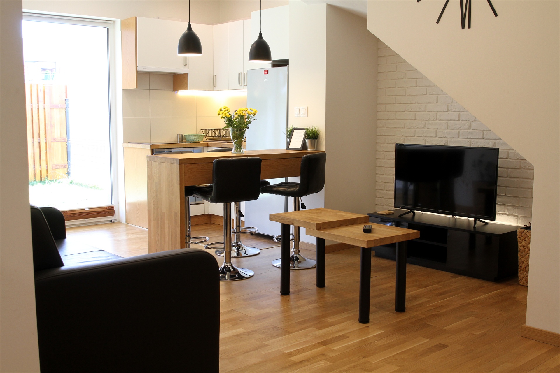 Kawalerka z drewnianym barem i dwoma czarnymi hokerami, wazon z żółtymi kwiatami i telewizor przy ścianie