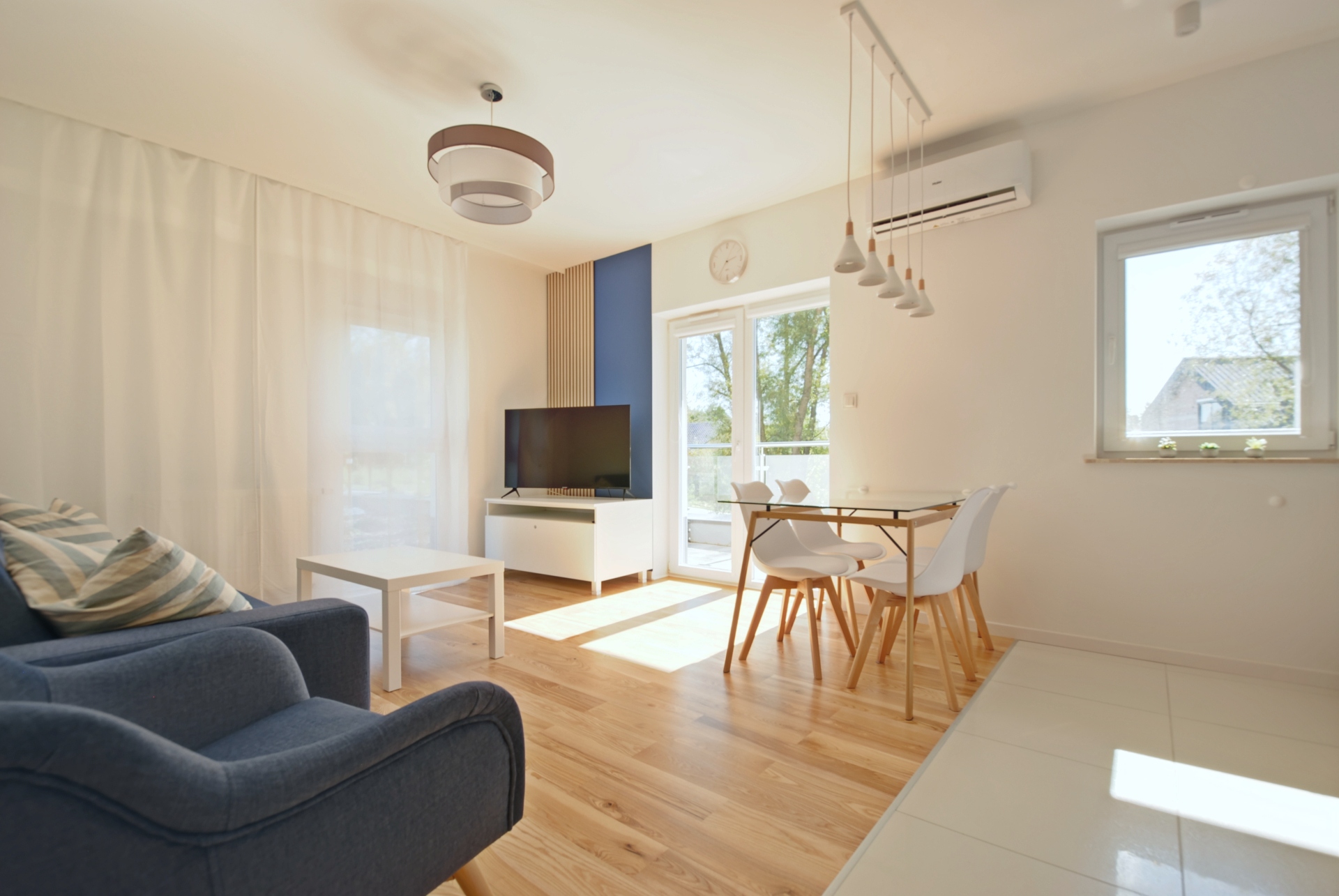 Jasny salon z niebieską sofą, białym stolikiem, stołem jadalnym, telewizorem i dużymi drzwiami balkonowymi