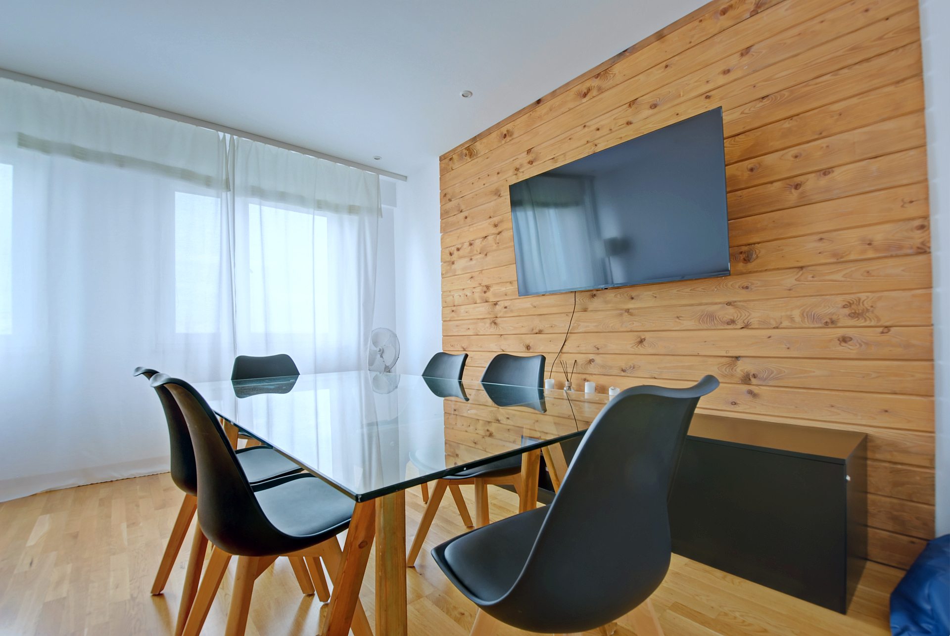 Szklany stół z czarnymi krzesłami przed drewnianą ścianą z zawieszonym telewizorem i jasnymi firanami